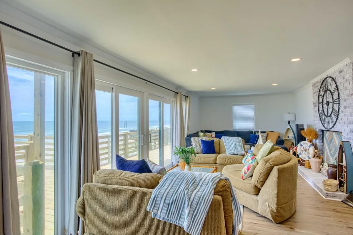 A cozy living area boasting a large, plush sectional sofa arranged in front of expansive windows that provide sweeping views of the Atlantic. Sunlight fills the room, highlighting a decorative wall clock and inviting nooks perfect for family gatherings or quiet reading. The ocean’s blue expanse is visible just beyond the glass, creating a seamless connection between the interior and the outdoors.