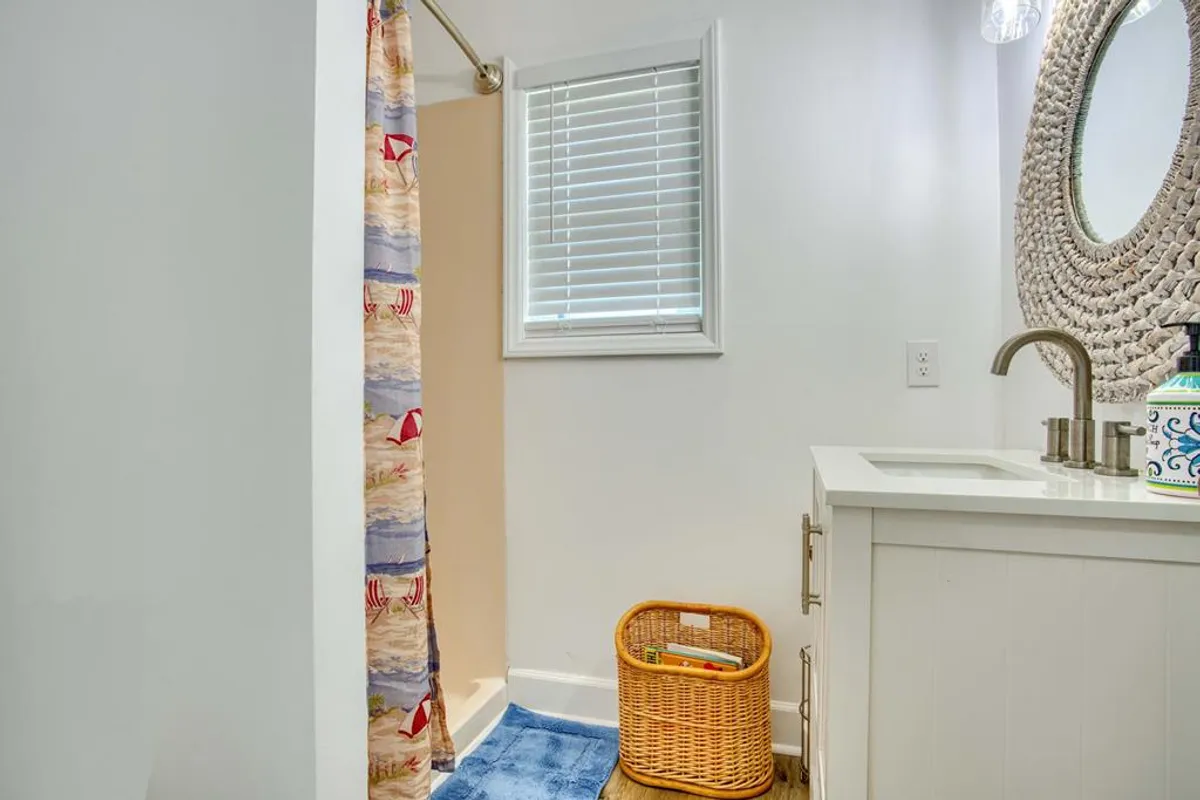 An inviting interior bathroom with a shower curtain featuring a beach motif, woven mirror, and decorative sink area. The space is bright and cheerful, evoking the feeling of freshening up after a beach day.