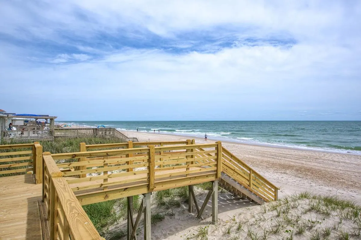 A broad wooden boardwalk stretches out towards the sandy beach, with the shimmering ocean visible in the background. People stroll along the boardwalk and beach, soaking up the relaxed, vacation atmosphere. The scene conveys the carefree spirit of Surf City, with open access to the water and opportunities for family outings, morning walks, or watching surfers.