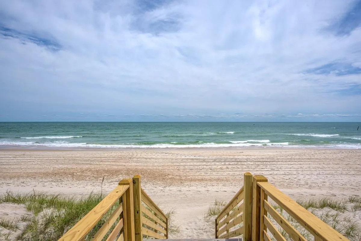 A wooden staircase descends to the sandy shoreline with gentle ocean waves in view. The scene captures the peaceful atmosphere of the beach at sunrise or sunset, perfect for making family memories.