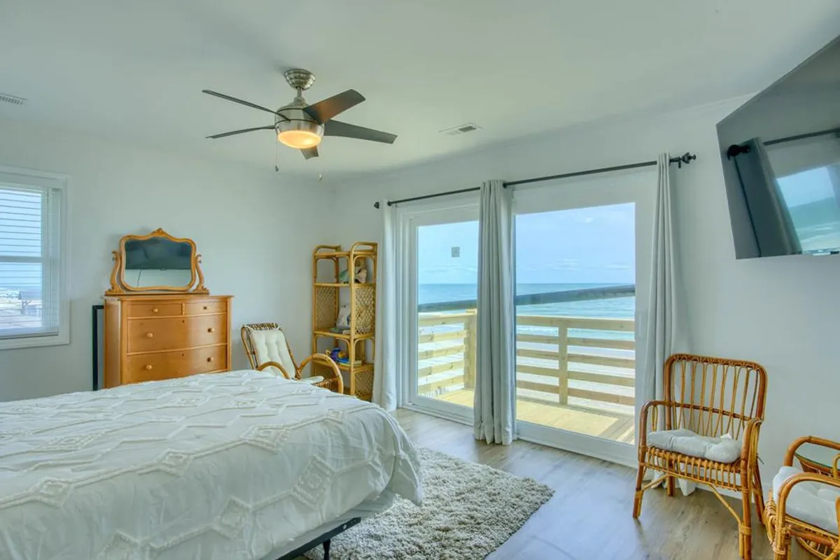 A bright bedroom with crisp white bedding, large windows looking out over the ocean, a wooden dresser, and rattan chairs. Natural light fills the room, highlighting both the cozy sleeping area and the stunning water views.