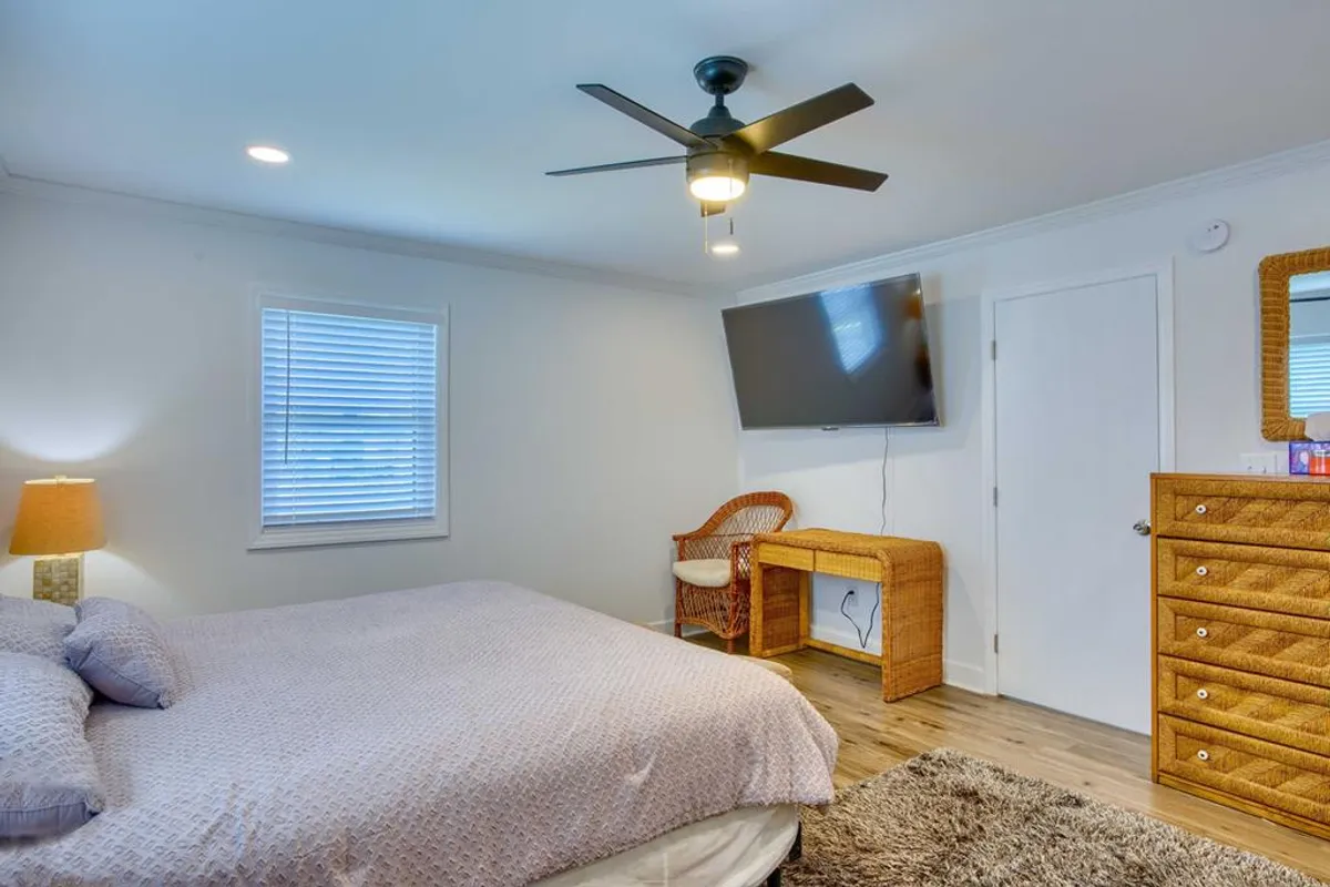 A serene guest bedroom with light purple bedding, a woven chair perfect for reading, a TV mounted on the wall, and a wooden dresser with a wicker design. The ambiance is peaceful and inviting, with ample natural light adding to the sense of a private sanctuary.