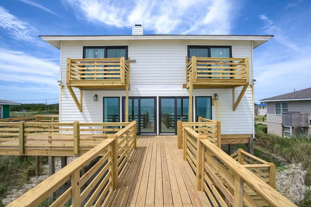 A tranquil wooden deck features comfortable seating and large glass doors opening to expansive ocean views. This inviting exterior space is ideal for relaxing, sipping coffee, or sharing stories after a day of exploring Surf City. The balconies and open design suggest easy movement between indoors and outdoors.