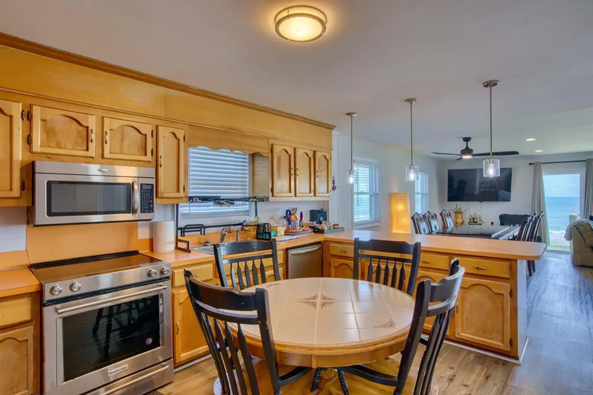 An open-concept kitchen and dining area filled with light from large windows. Warm wooden cabinets, a round dining table, and stainless steel appliances invite gatherings for shared meals, while the ocean view provides a picturesque backdrop. This space is perfect for family feasts and casual breakfasts alike.