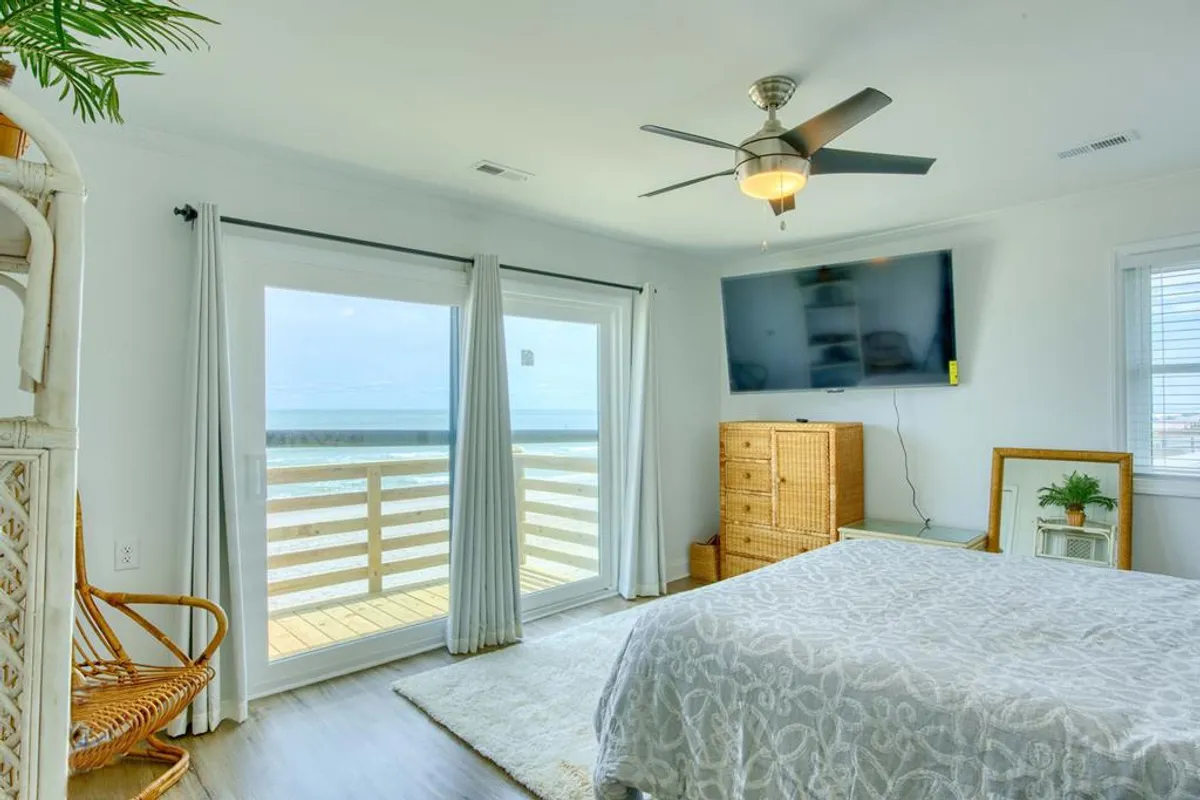 A tranquil bedroom featuring a comfortable bed, large balcony doors opening to a private oceanfront view, a ceiling fan, and a TV. Soft natural light fills the space, inviting relaxation and a sense of retreat. The balcony hints at the soothing sound of waves and a perfect spot for morning coffee or sunrise watching.