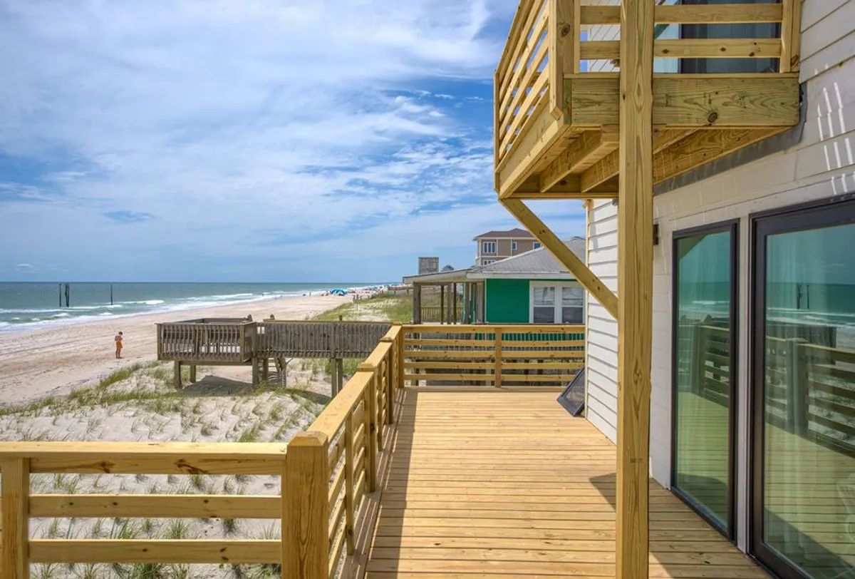 A wide deck overlooks the beach and ocean, with rolling waves under a cloudy sky. The deck is perfect for sunbathing, relaxing, or gathering for a sunset barbecue, with easy access to the sand below. The scene hints at group and family activities, tying in with Surf City's social and recreational options.