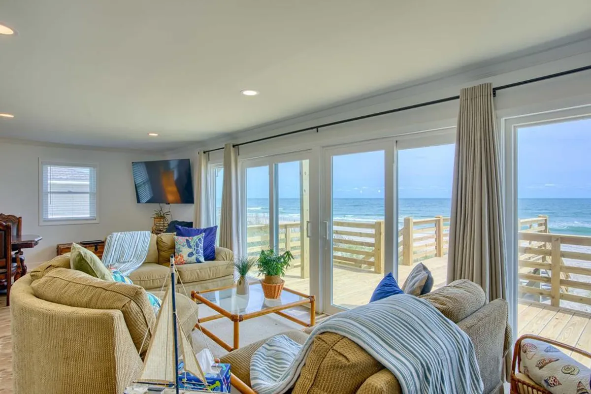 The image captures a spacious, sunlit living room with plush, inviting seating positioned to take in sweeping ocean views through large windows. The coffee table is decorated with tasteful coastal accents, and the tranquil atmosphere is perfect for family relaxation or quiet moments watching the waves.