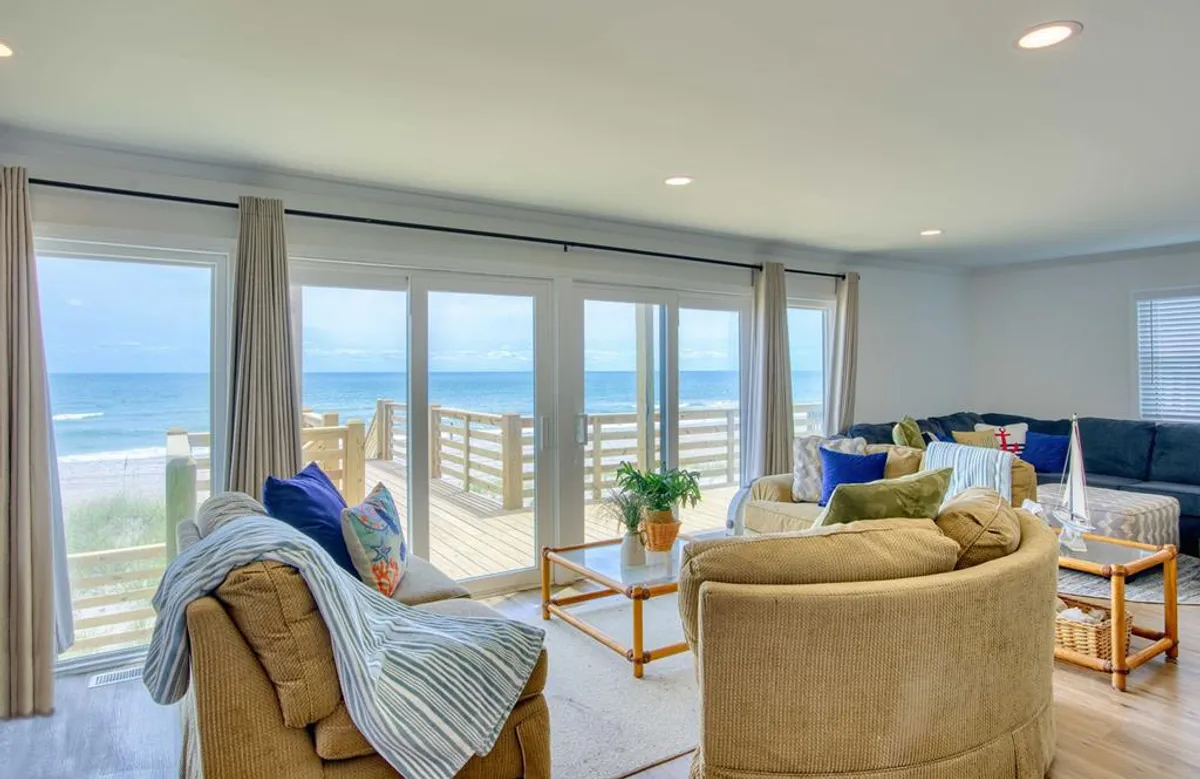 A bright, inviting space with two sofas oriented towards a large set of windows, providing unbroken views of the ocean. The room features deck access, a coffee table, and greenery, blending indoor comfort with the allure of the outdoors. Natural light floods the space, making it ideal for relaxation and social connection.