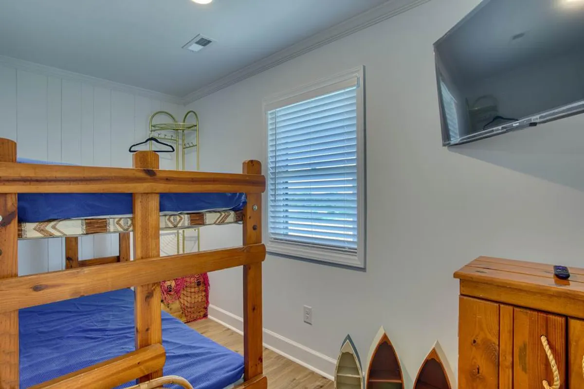 A cheerful bunkroom featuring a bunk bed, wooden dresser, wall-mounted TV, and playful surfboard decorations. The room is bright and welcoming, perfect for kids or friends to share.